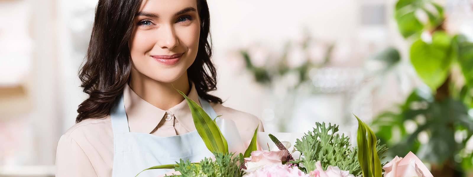 Smiling young florist holding a bouquet of fresh pink roses and greenery in a bright flower shop. 微笑花藝師手捧粉紅玫瑰與綠葉花束，背景明亮溫馨的花店內。